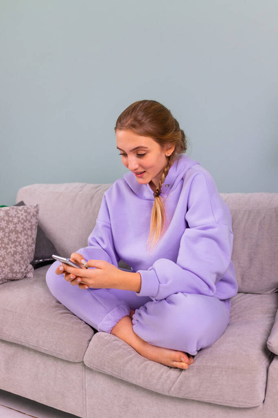 Happy smiling teenage girl with her smartphone browsing social newtwork, communicating relaxed sitting on a sofa at home. Distance online education, study from home concept. - Фото, зображення