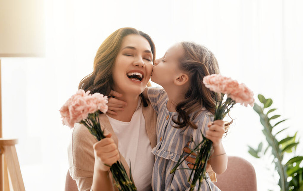 Happy day! Child daughter is congratulating mother and giving her flowers. Mum and girl smiling and hugging. Family holiday and togetherness. - Фото, изображение