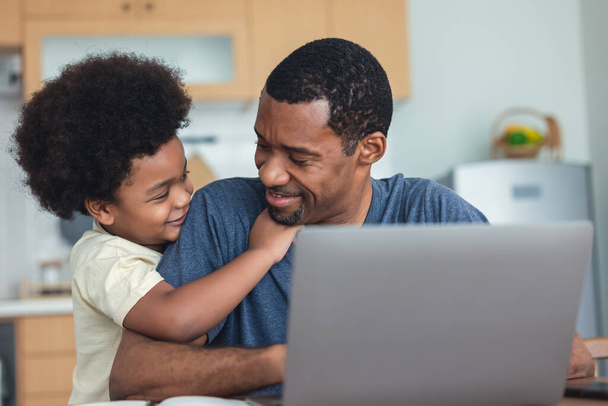 Porträt eines süßen afrikanischen Jungen, der seinen hübschen Vater umarmt, der am Laptop arbeitet, während er zu Hause am Küchentisch sitzt. Afroamerikanische Familie verbringt Zeit zu Hause mit Kind. - Foto, Bild