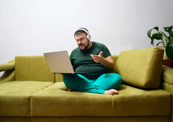 serious bearded male with laptop in wireless headphones and casual clothes sitting alone on soft couch, listening to favorite music  - Фото, зображення