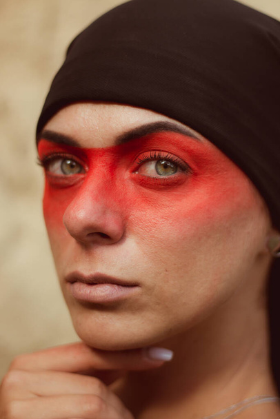 Portrait of a young beautiful woman with makeup and face art in American tribal or Middle Eastern style. Ethnic image, red eyes and black headscarf. Close-up. - Foto, imagen
