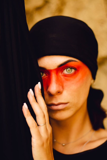 Portrait of a young beautiful woman with makeup and face art in American tribal or Middle Eastern style. Ethnic image, red eyes and black headscarf. Close-up. - Foto, Imagem