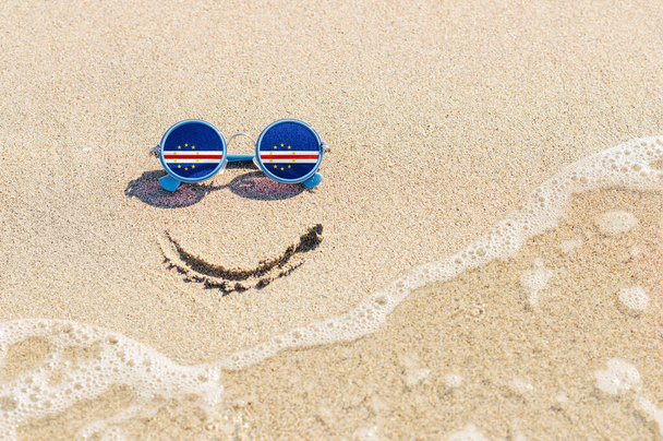 Ένα ζωγραφισμένο χαμόγελο στην άμμο και γυαλιά ηλίου με τη σημαία του Πράσινου Ακρωτηρίου. Η έννοια των θετικών και επιτυχημένων διακοπών στο θέρετρο του Πράσινου Ακρωτηρίου. - Φωτογραφία, εικόνα