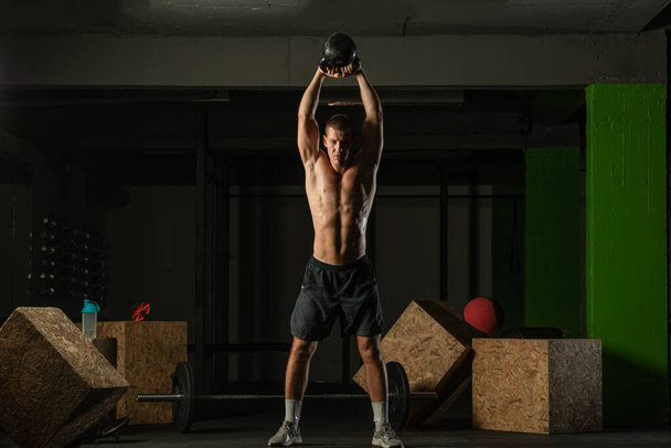 Plná fotografie pohledného muže s nahým trupem cvičení s kettlebell na tmavém pozadí - Fotografie, Obrázek