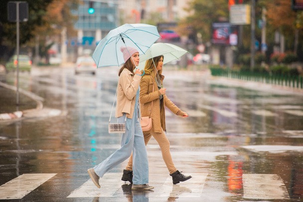 Atrakcyjne i młode siostry przechodzą razem przez ulicę podczas deszczu. - Zdjęcie, obraz