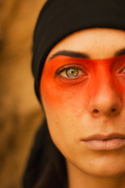 Portrait of a young beautiful woman with makeup and face art in American tribal or Middle Eastern style. Ethnic image, red eyes and black headscarf. Close-up. - Zdjęcie, obraz