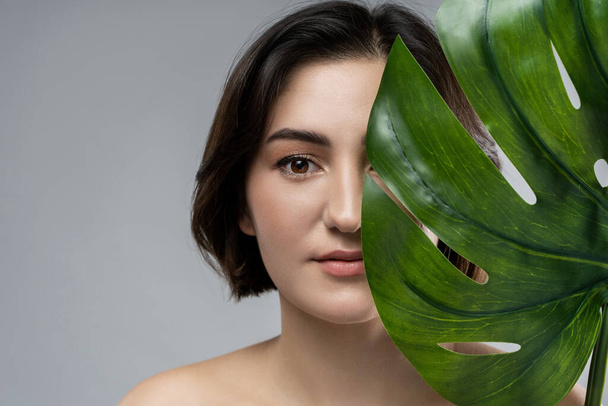 Portrait of young beautiful brunette woman with a monstera deliciosa tropical leaf on gray background - Photo, Image