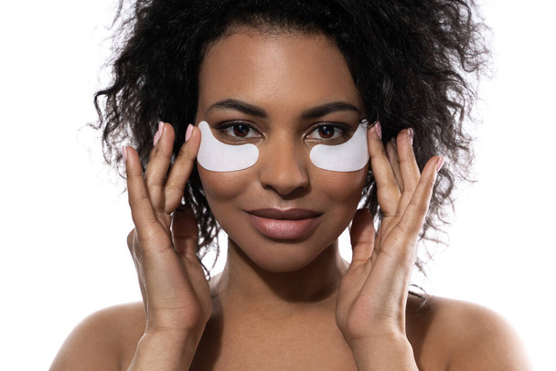 Beautiful black woman with a smooth skin applying adhesive under-eyes patches for dark circles on white background - Foto, imagen