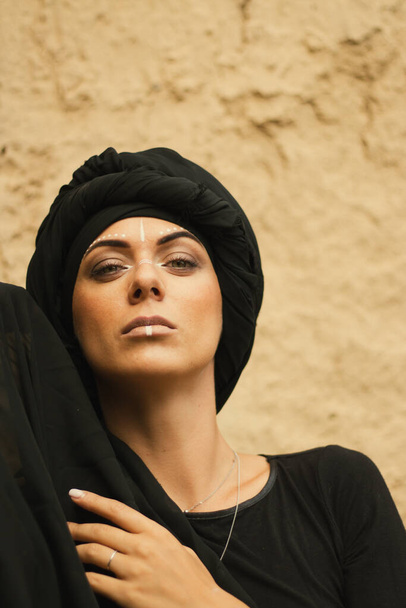 Portrait of a young beautiful woman with makeup and white face art ornament in tribal or middle eastern style dressed in a black hijab. Ethnic image Close-up on the background of a sandy wall. - Фото, зображення