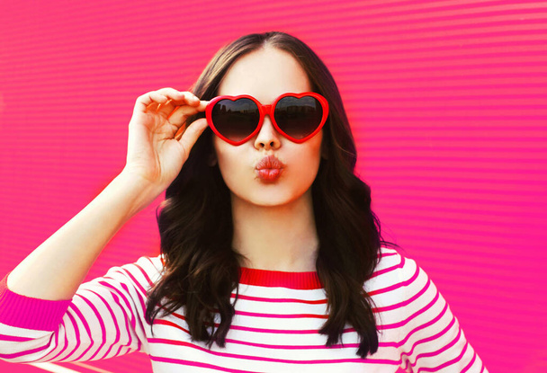 Portrait of beautiful young brunette woman blowing her lips wearing red heart shaped sunglasses on pink background - Fotografie, Obrázek