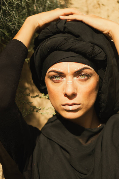 Portrait of a young beautiful woman with makeup and white face art ornament in tribal or middle eastern style dressed in a black hijab. Ethnic image Close-up on the background of a sandy wall. - Fotografie, Obrázek