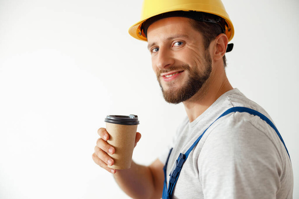 Портрет веселого мастера в форме и каске, улыбающегося в камеру во время кофе-брейка - Фото, изображение