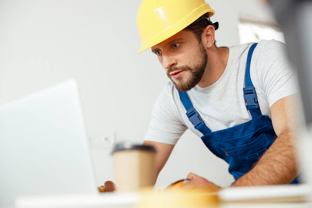 Handyman enfocado en uniforme y sombrero duro usando el ordenador portátil mientras que comienza trabajos de renovación en el interior - Foto, imagen