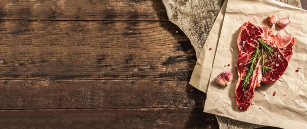 Κλασσική μπριζόλα T-bone με φρέσκο δενδρολίβανο, σκόρδο και μπαχαρικά. Ένα μοντέρνο σκληρό φως, σκούρα σκιά, μοντέρνο περίπτερο, ξύλινο υπόβαθρο, επίπεδη lay. Ξύλινο φόντο, ρουστίκ στυλ, μορφή banner - Φωτογραφία, εικόνα