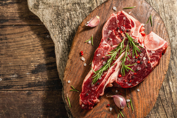 Classic T-bone steak with fresh rosemary, garlic and spices. A modern hard light, dark shadow, trendy stand, wooden background, flat lay. Wooden background, rustic style, top view - 写真・画像