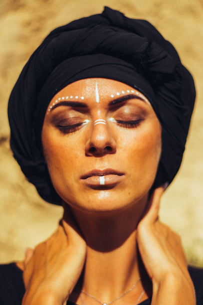Retrato de una joven hermosa mujer con maquillaje y adorno de arte de cara blanca en estilo tribal o de Oriente Medio vestida con un hiyab negro. Imagen étnica Primer plano sobre el fondo de una pared arenosa. - Foto, Imagen