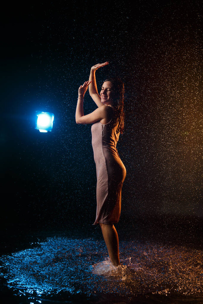 Beautiful sexy girl with drops of water on her face in splashes illuminated by an orange light against a dark background in the studio. - Foto, immagini