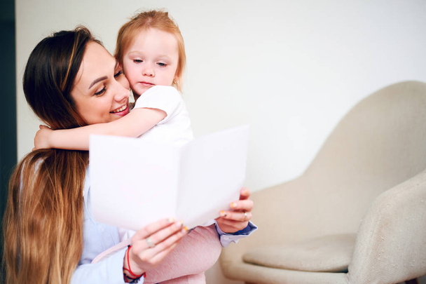 С Днем Матери. Счастливая улыбающаяся мама с маленькой дочкой читает праздничную открытку, сидя на полу - Фото, изображение