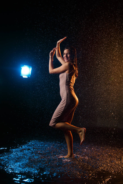 Beautiful sexy girl with drops of water on her face in splashes illuminated by an orange light against a dark background in the studio. - Foto, immagini