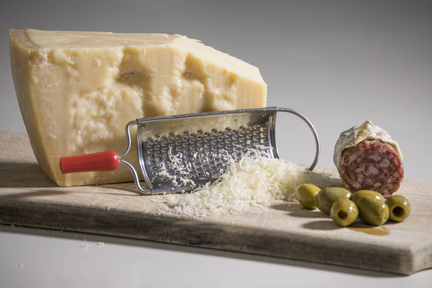 Een persoon raspt parmezaanse kaas op een houten snijplank met olijven en salami. - Foto, afbeelding