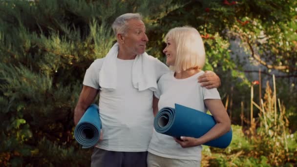 Χαμογελώντας ηλικιωμένο ζευγάρι με στρώματα γιόγκα στέκεται έξω, έτοιμο για προπόνηση - Πλάνα, βίντεο