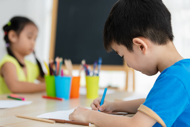 Τα παιδιά από την Ασία χρησιμοποιούν χρωματιστό μολύβι και στυλό για να ζωγραφίσουν χαρούμενα εικόνες στο τραπέζι της τάξης πίσω στο σχολείο. - Φωτογραφία, εικόνα