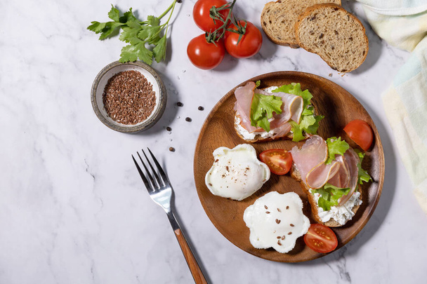 Easter breakfast or brunch. Delicious breakfast or snack - poached egg and cream cheese toast whole grain rye bread, prosciutto, arugula on a marble tabletop. Top view flat lay. Copy space.  - Фото, изображение