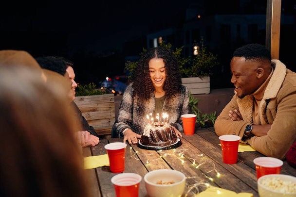 Gruppo di diversi giovani adulti che festeggiano il compleanno femminile biennale, seduti alla festa sul tetto alla moda che celebra la festeggiata con torta. - Foto, immagini