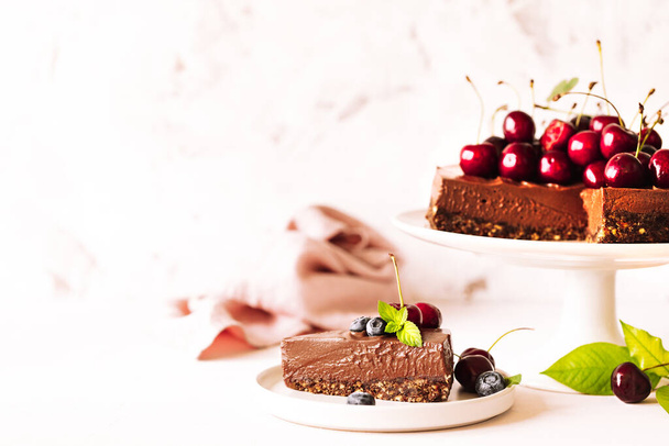 Un pezzo di torta al cioccolato decorata con ciliegie fresche, mirtilli e un'intera torta su uno stand di ceramica su uno sfondo beige. Orizzonte. - Foto, immagini