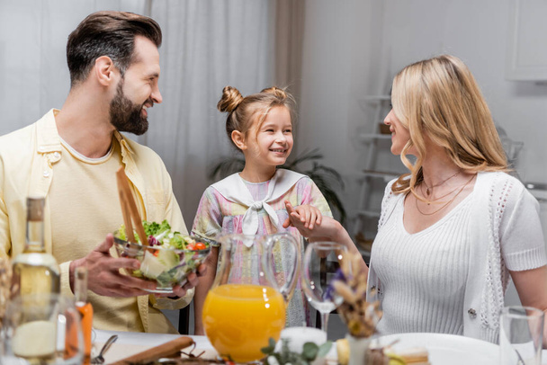 szczęśliwy człowiek trzyma sałatkę warzywną w pobliżu żony i córki trzymając się za ręce podczas wielkanocnego obiadu - Zdjęcie, obraz