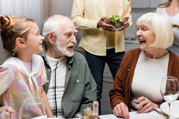 senior woman laughing near husband and granddaughter during family dinner - Photo, Image