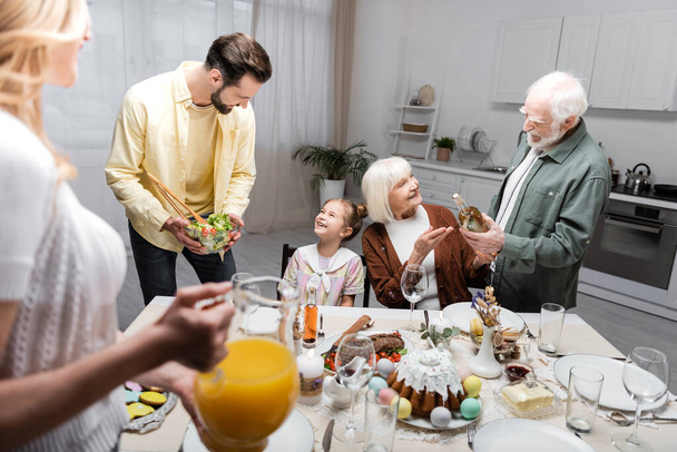 men holding bottle of wine and vegetable salad during easter dinner with family - Фото, зображення