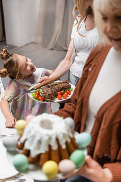 gelukkig meisje glimlachen in de buurt van moeder en wazig oma met Pasen taart en vlees met groenten - Foto, afbeelding