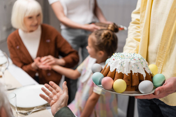 man holding delicious easter cake and painted eggs near blurred family - Foto, Imagen
