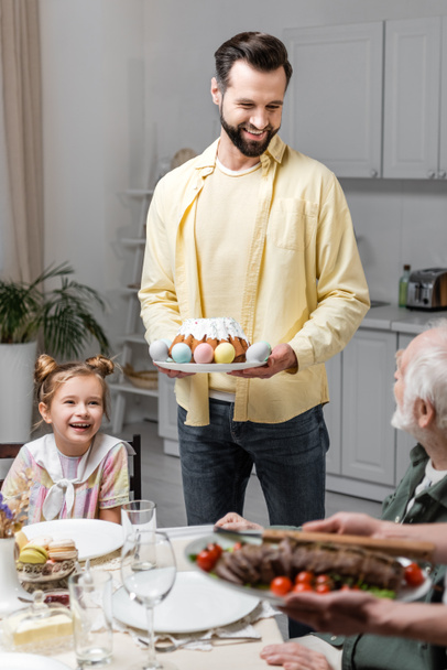 smiling man holding easter cake and painted eggs near family during festive dinner - Photo, Image