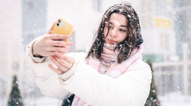 雪の降る冬の天候の中でスマートフォンを屋外で10代の女の子の肖像画. - 写真・画像