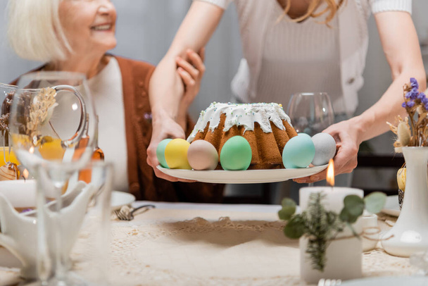 osittainen näkymä nainen tilalla pääsiäiskakku ja maalattu munia lähellä vanhempi äiti aikana juhlapäivällinen - Valokuva, kuva