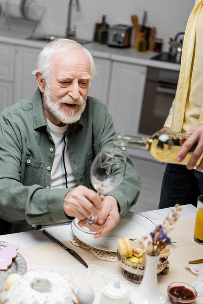 senior man holding glass near adult son pouring wine during easter dinner - Foto, Imagen