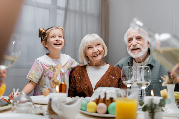 cheerful girl smiling near happy grandparents and festive dinner on table - Foto, imagen
