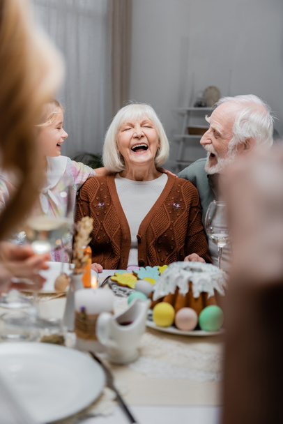 пожилая женщина смеется с закрытыми глазами рядом с семьей и пасхальный ужин на размытом переднем плане - Фото, изображение
