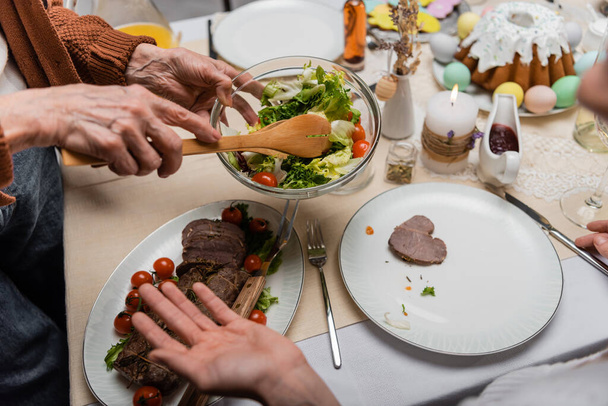 gedeeltelijk zicht op oudere vrouw die groentesalade in de buurt van vlees op feestelijke tafel houdt - Foto, afbeelding