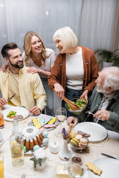 senior woman holding bowl with vegetable salad near happy family  - Foto, Imagen