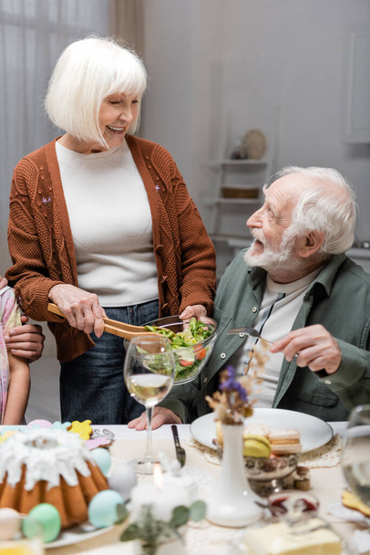 senior woman holding vegetable salad near happy husband during easter dinner - Foto, Imagen