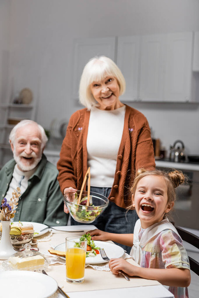 seniorka trzyma sałatkę warzywną w pobliżu wesołego męża i śmiejącej się wnuczki - Zdjęcie, obraz