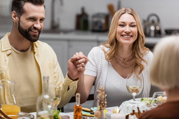 alegre pareja cogida de la mano cerca de la mesa servida con cena de Pascua en primer plano borroso - Foto, imagen