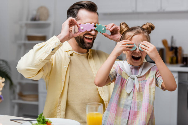 cheerful man with daughter covering eyes with easter eggs and cookies while having fun at home - Fotó, kép