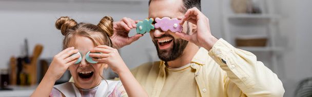 śmiejący się ojciec i córka przykrywający oczy wielkanocnymi ciasteczkami i malowanymi jajkami podczas zabawy w domu, sztandar - Zdjęcie, obraz