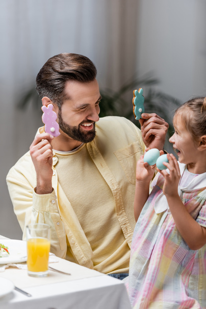 podekscytowana dziewczyna z tatą bawiąc się z malowanymi jajkami i tradycyjnymi ciasteczkami podczas Wielkanocy - Zdjęcie, obraz