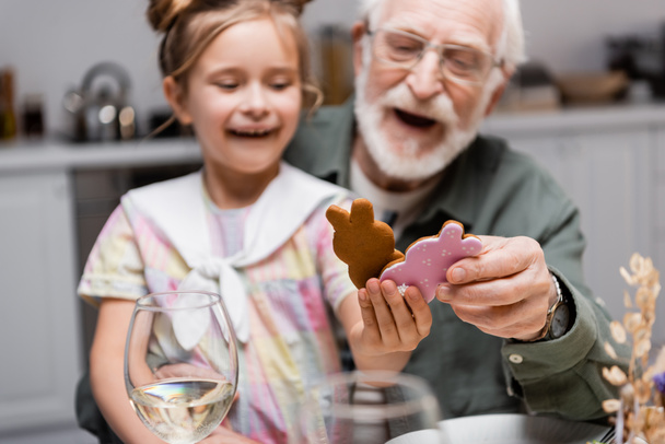 erstaunt Mädchen mit Opa hält Osterkekse während Feier zu Hause, verschwommener Hintergrund - Foto, Bild