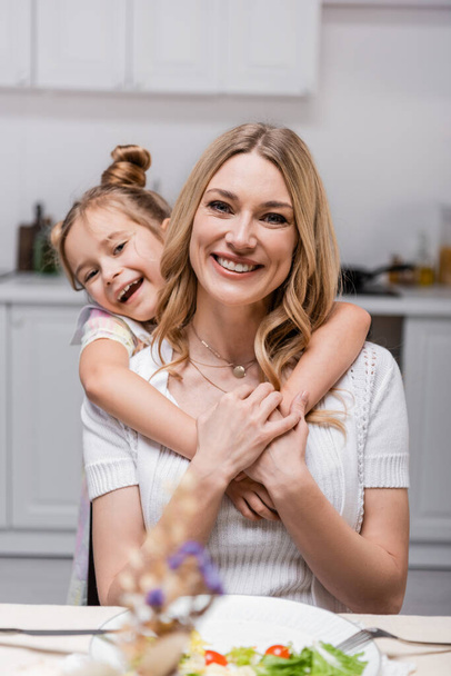 cheerful girl embracing happy mother while looking at camera near served dinner - Photo, Image